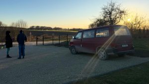 Cilvēku grupa stāv pie sarkana mikroautobusa lauku pagalmā, fonā redzams saulriets un kaili koki.