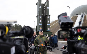Vācijas militārpersona sniedz preses brīfingu pie raķešu sistēmas, žurnālistu kamerām un mikrofonu ielenkumā.
