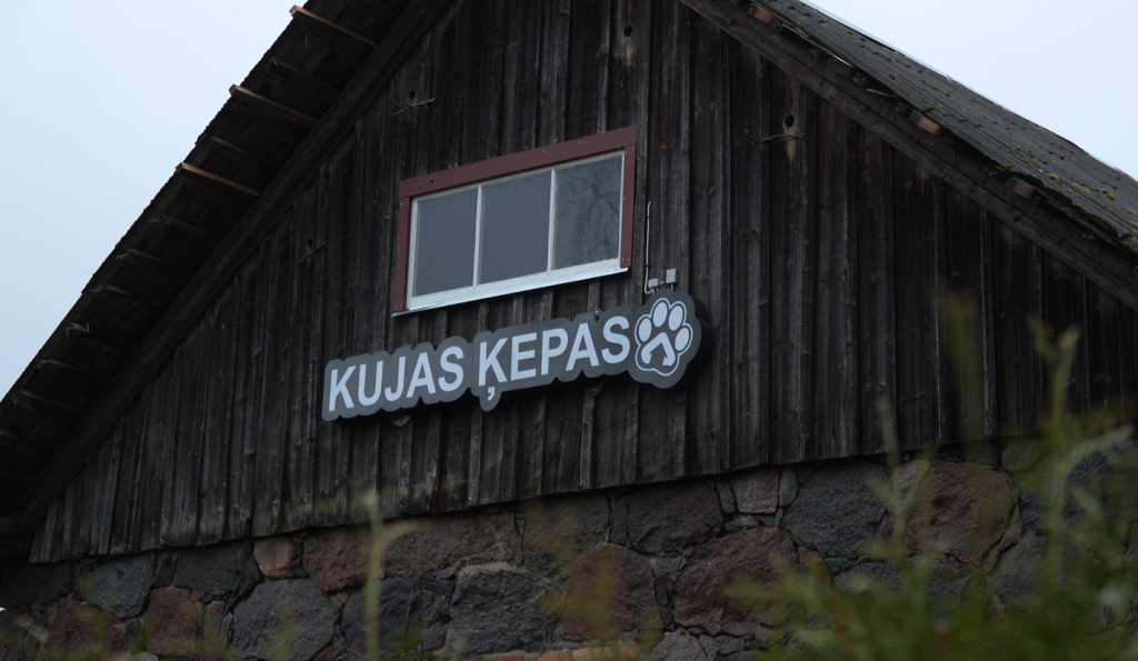 Koka ēkas zelminis ar izkārtni "Kujas Ķepas" un ķepas simbolu virs akmens mūra sienas.