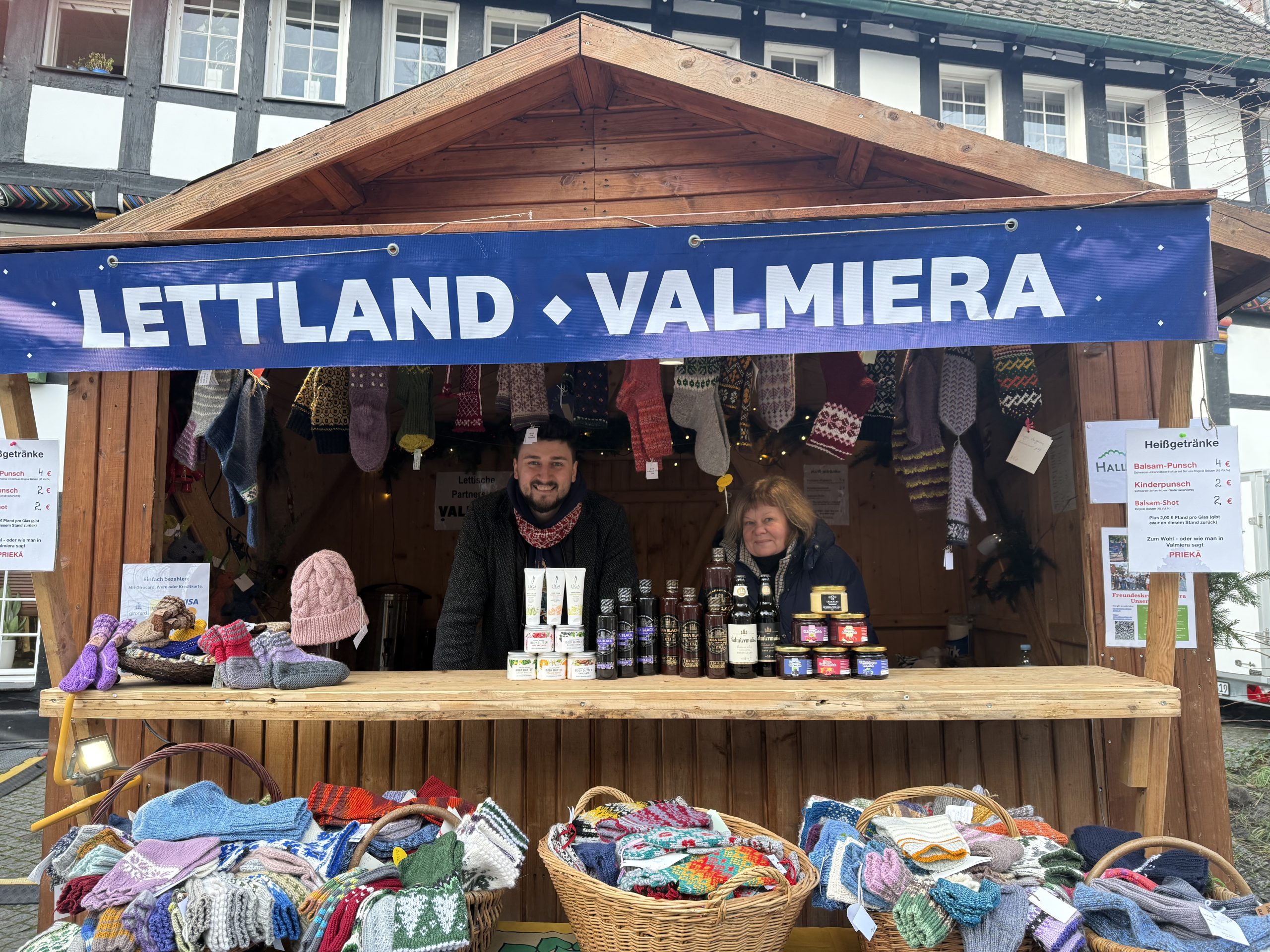 Valmieras amatnieku namiņš Vācijas Ziemassvētku tirdziņā ar rokdarbiem, pārtikas produktiem un diviem pārstāvjiem pie letes.