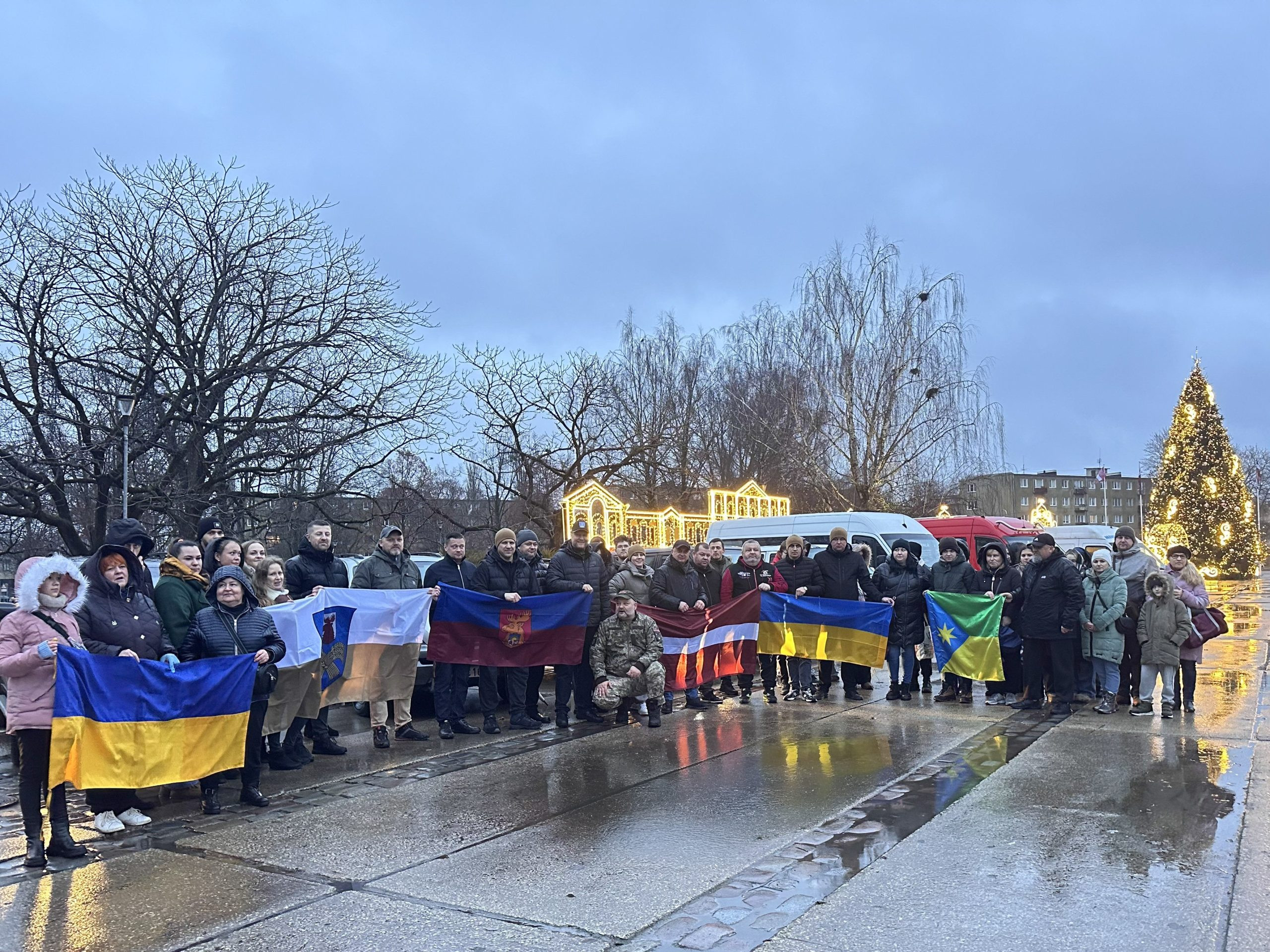 Cilvēku grupa ar Latvijas, Ukrainas un pilsētu karogiem pulcējas svinīgā pasākumā pie izgaismotas Ziemassvētku egles.