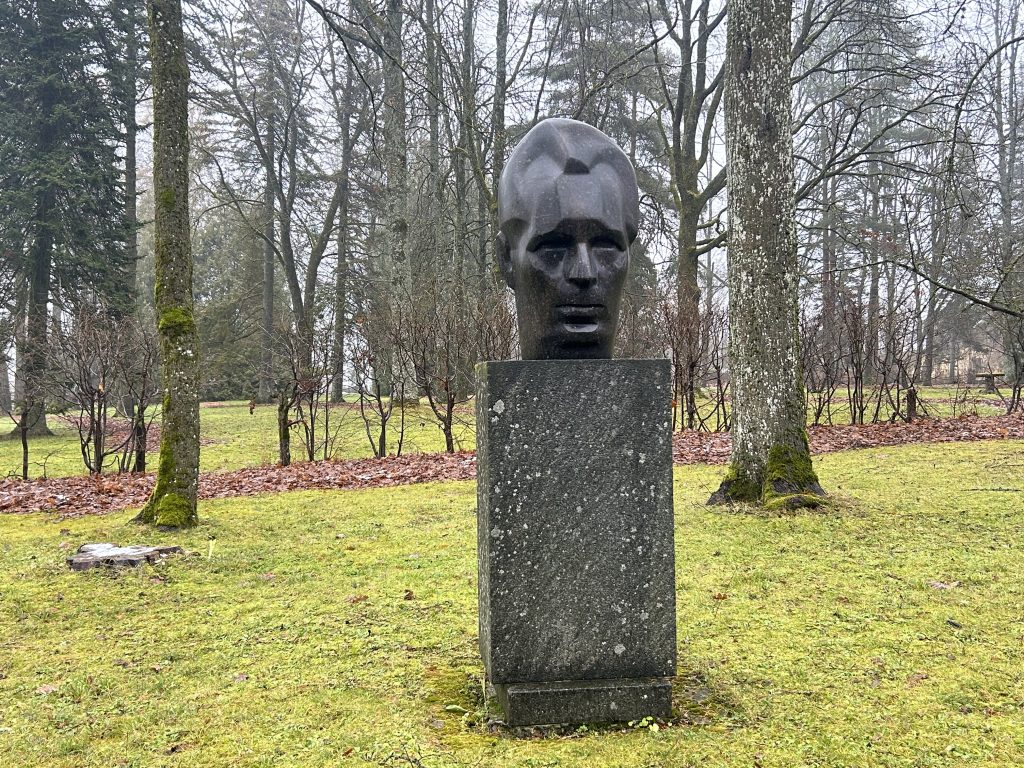 Tuvplāns uz tumši pelēku vīrieša galvas skulptūru (Andrejs Upītis) uz augsta, monolīta pjedestāla zaļā parkā.