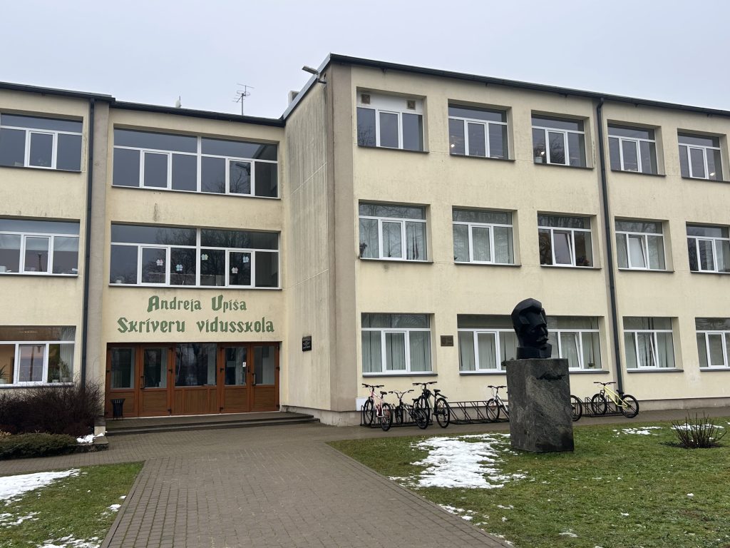 Gaiši dzeltena divstāvu skolas ēka ar lieliem logiem. Pie ieejas uzraksts zaļā krāsā: "Andreja Upīša Skrīveru vidusskola".