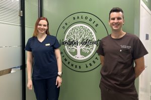 Divi cilvēki, vīrietis un sieviete, medicīnas tērpos stāv pie zaļas sienas ar baltu koka logotipu: "Dr. Balodis. Veselības doktorāts. Ģimenes ārsts."