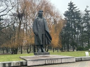 Bronzas krāsas statuja, kas attēlo vīrieti garā mētelī (Andrejs Upīts) stāvam uz zema, bet masīva pjedestāla parkā.