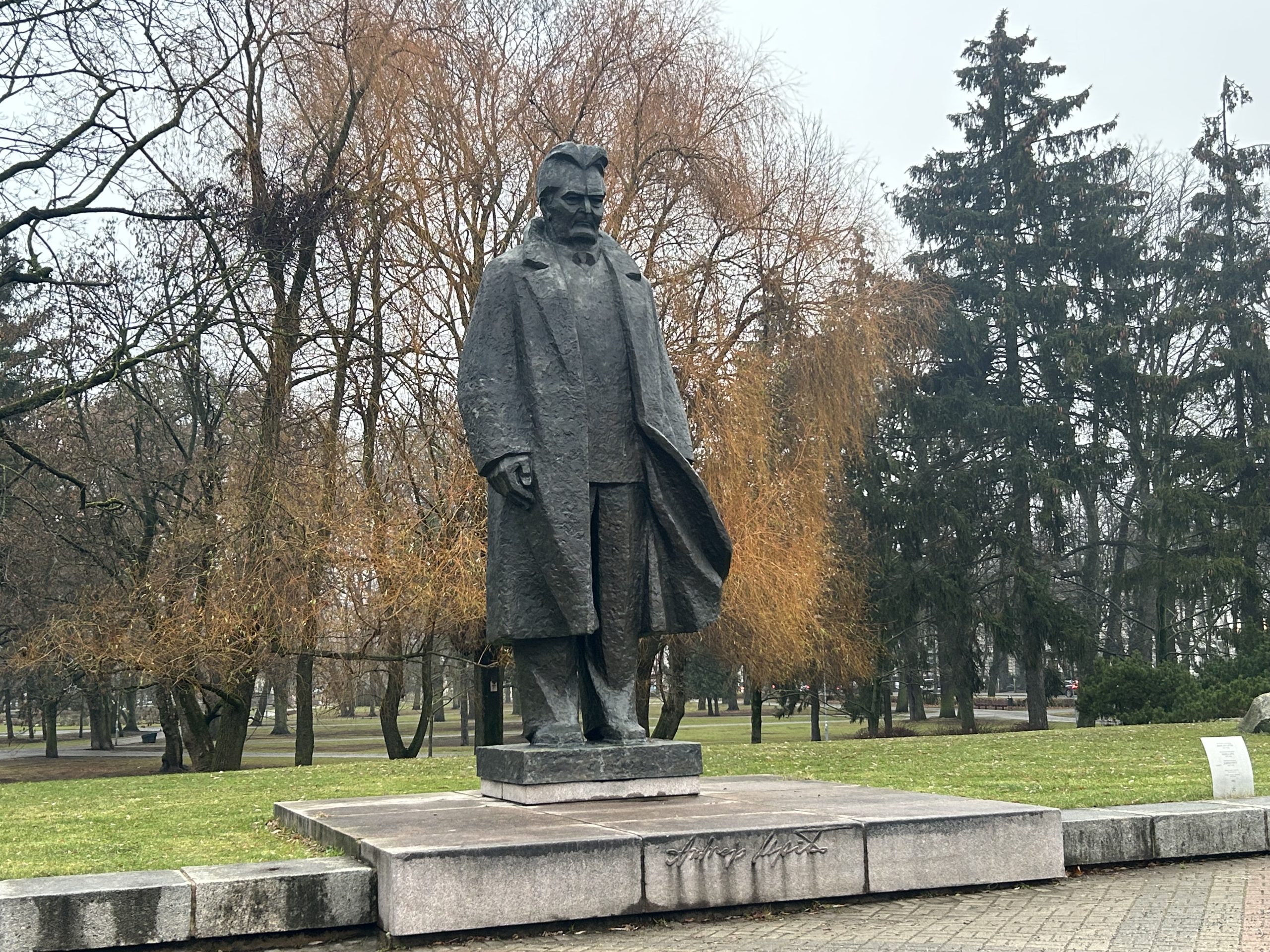 Bronzas krāsas statuja, kas attēlo vīrieti garā mētelī (Andrejs Upīts) stāvam uz zema, bet masīva pjedestāla parkā.