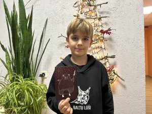 Bērns tur rokās Ziemassvētku pastkarti ar zīmētu lāci, stāvot pie svētku dekorācijas un telpaugiem.