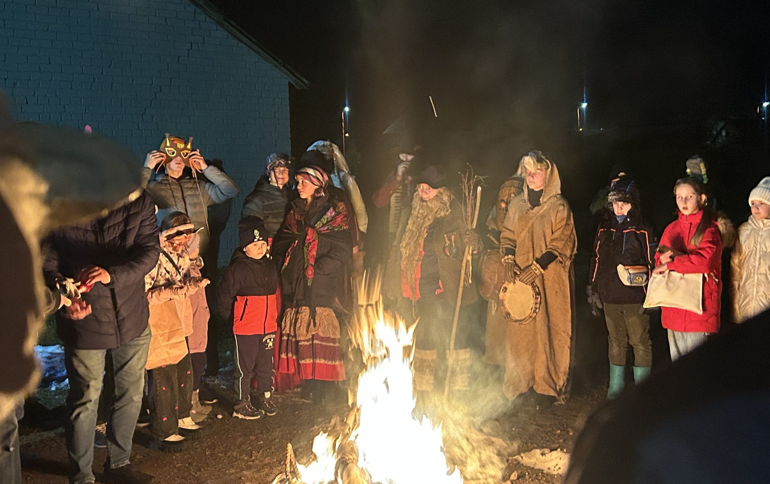 Cilvēku grupa, tostarp bērni un pieaugušie maskās, stāv ap lielu ugunskuru tumsā.