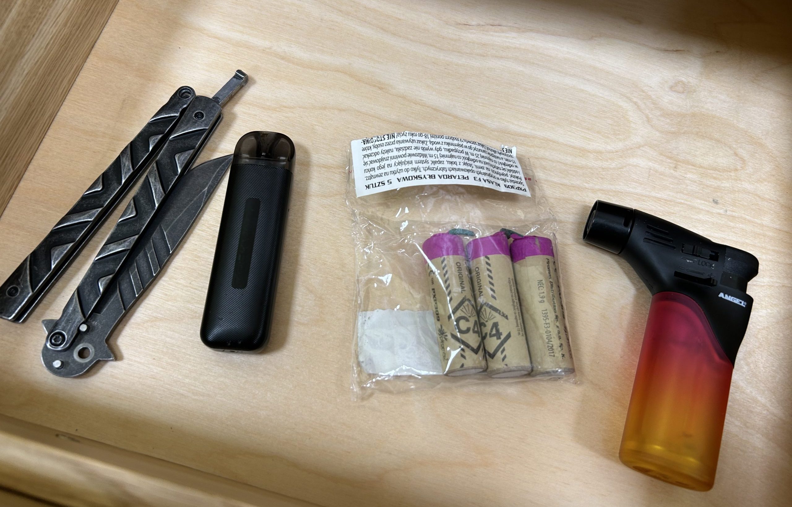 Atvilktnē redzami konfiscētie aizliegtie priekšmeti: saliekamais nazis, e-cigarete, petaržu iepakojums un gāzes šķiltava.
