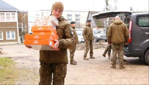 Zemessardze uniformā nes vairākas oranžas dāvanu kastes.