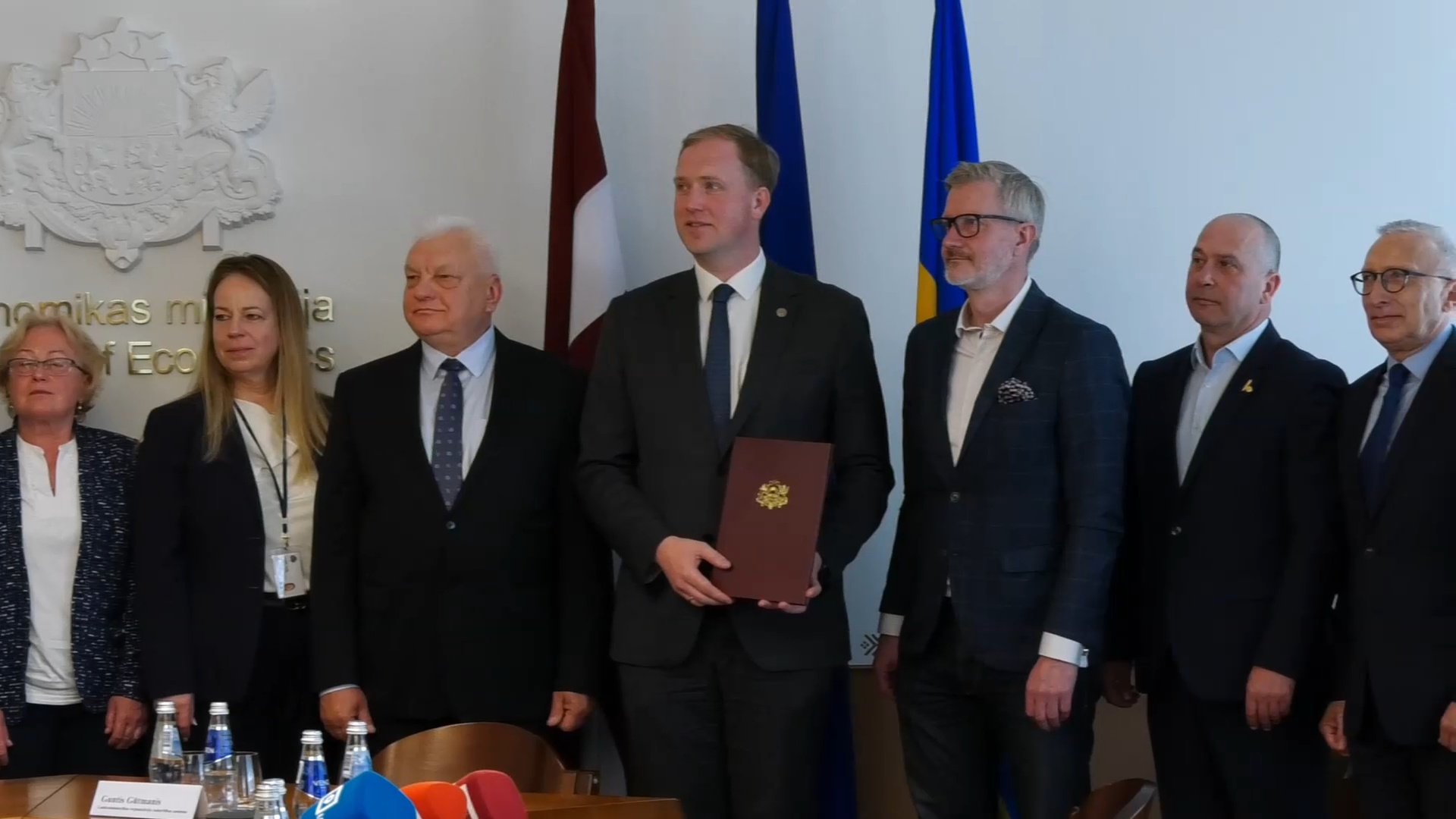 Septiņi cilvēki uzvalkos un lietišķā apģērbā stāv pie karogiem Ekonomikas ministrijā.