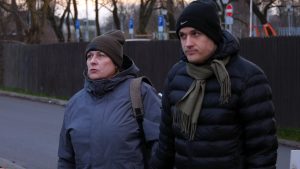 Divi cilvēki ziemas apģērbā stāv uz ielas, klausoties un vērojot apkārt notiekošo.