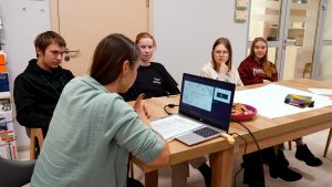 Četri jaunieši un viena pieaugušā sēž pie galda, skatoties un apspriežot informāciju portatīvajā datorā.
