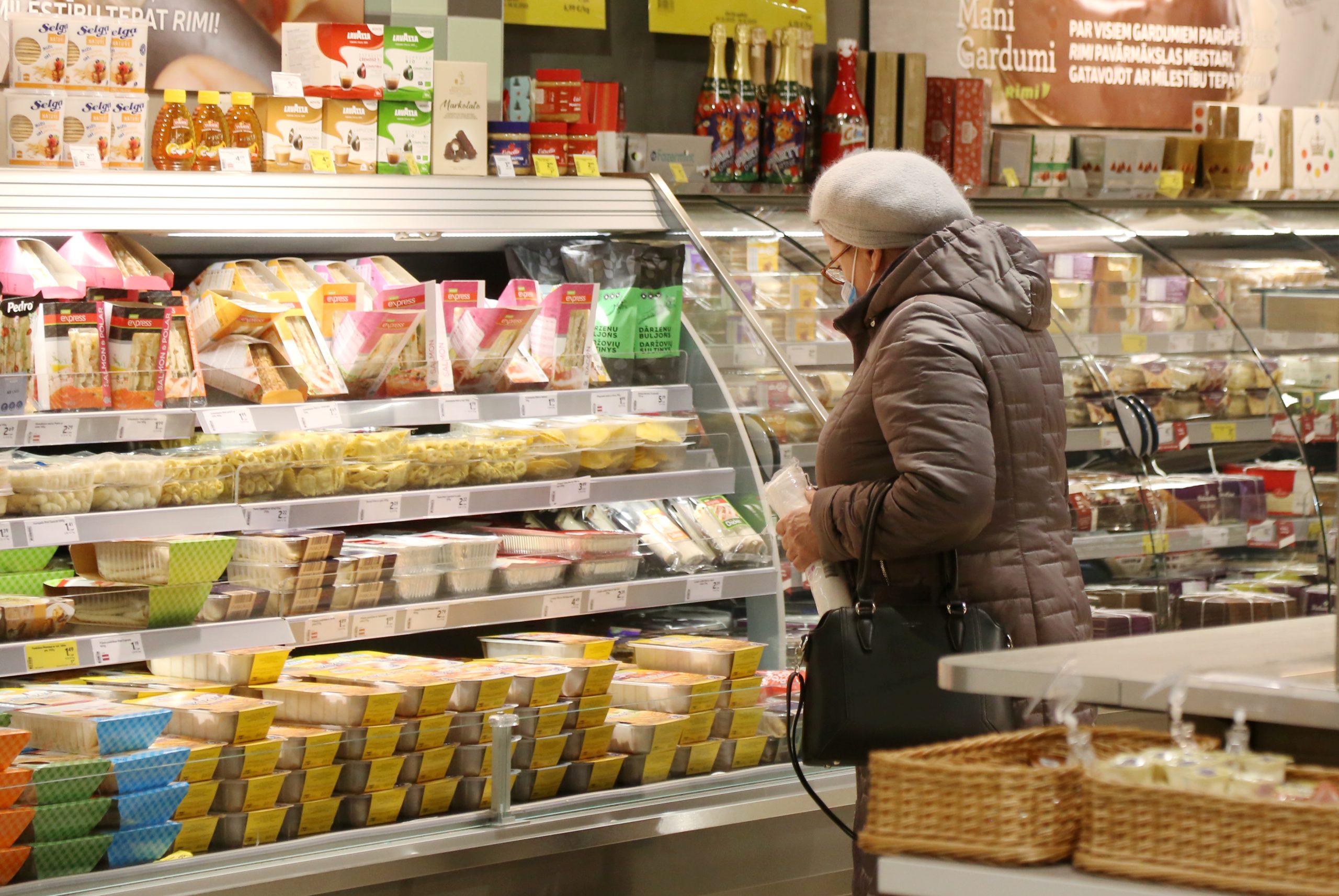 Pircēja pie pārtikas vitrīnas lielveikalā Latvijā, izvēloties produktus pie augstām cenām