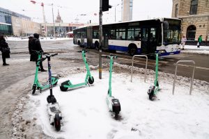 Vairāki koplietošanas elektroskrejriteņi novietoti sniegā pie Rīgas Centrālās stacijas laukuma; fonā brauc autobuss.