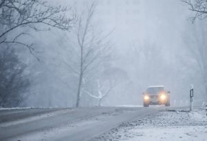 Automašīna ar ieslēgtām gaismām brauc pa sniegotu ceļu sliktas redzamības apstākļos