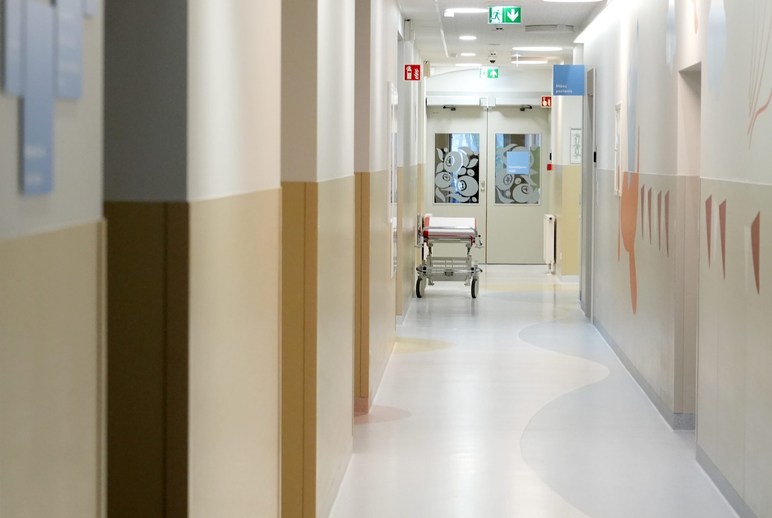 Tukšs Bērnu slimnīcas gaitenis ar medicīnisko gultu, skats uz nodaļas durvīm.
