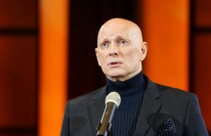 Jaunā Rīgas teātra mākslinieciskais vadītājs Alvis Hermanis runā mikrofonā uz oranža fona.