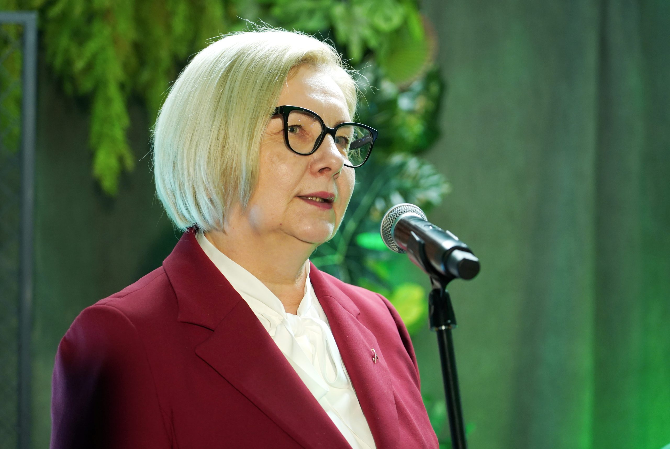 Rita Vectirāne, sieviete gados ar īsiem blondiem matiem un brillēm, uzstājas pie mikrofona sarkanā žaketē.