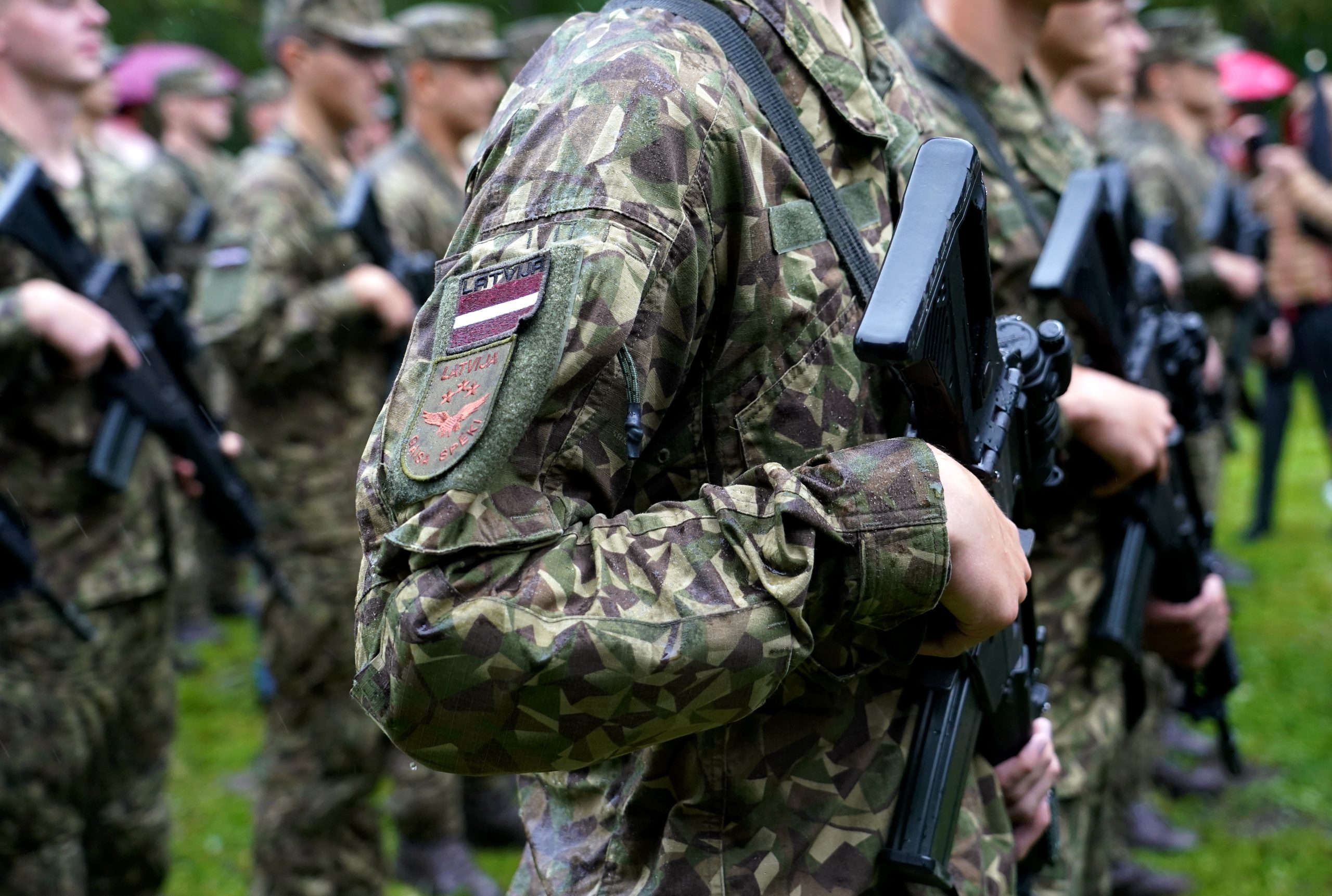 Latvijas karavīri maskēšanās formas tērpos stāv ierindā ar ieročiem.