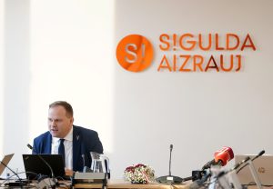Siguldas domes sēde, kurā pie galda runā amatpersona ar fonā redzamu uzrakstu “Sigulda aizrauj”.