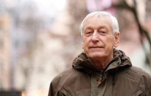 Tuvplāns: vecāks vīrietis ar sirmiem matiem, Jānis Kažociņš, brūnā ziemas jakā skatās tieši kamerā ar nelielu smaidu.