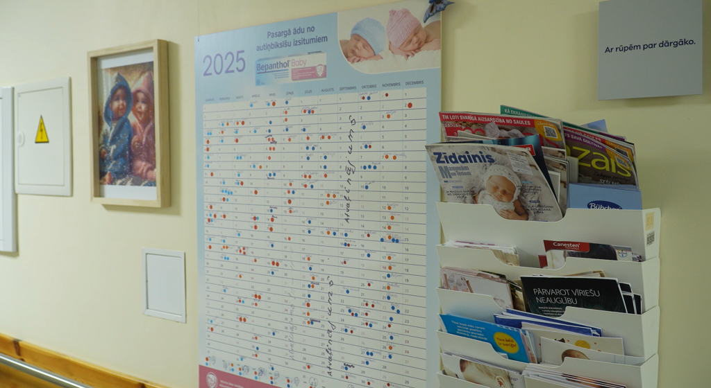Gaišā telpā uz sienas piestiprināts liels 2025. gada kalendārs ar atzīmēm, un blakus plauktiņš ar žurnāliem un informatīviem materiāliem.