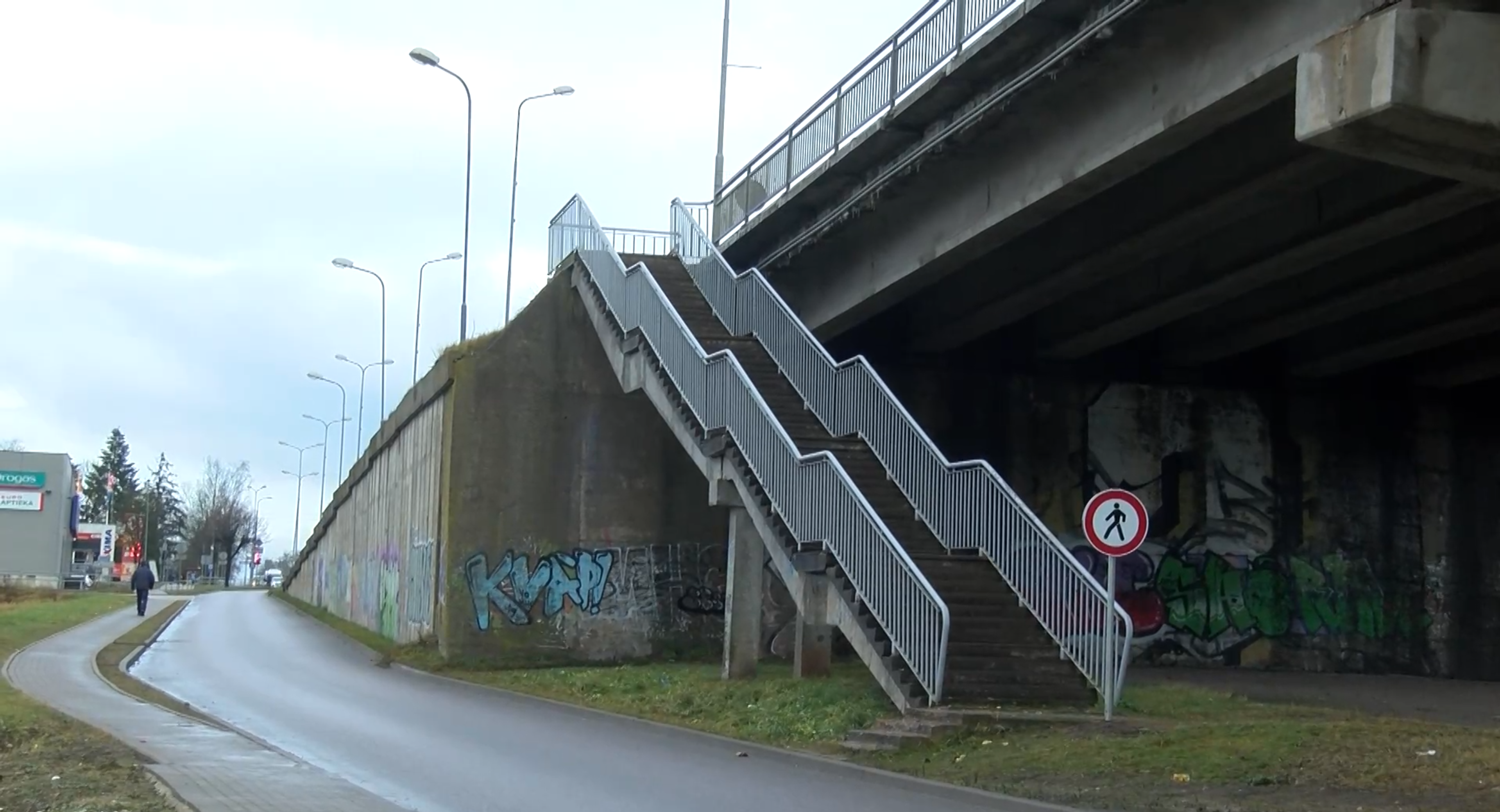 Gājēju kāpnes pie tilta pilsētvidē ar nolietotu infrastruktūru.