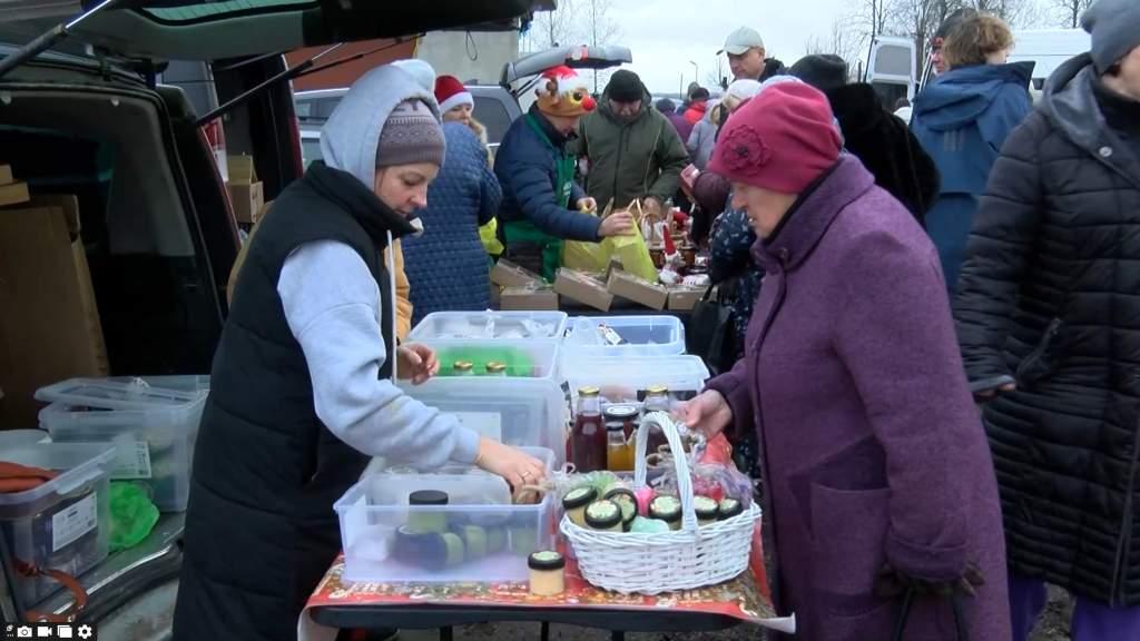 Tirgotāja rāda pircējai preces, kas izvietotas uz galda tirdziņā.