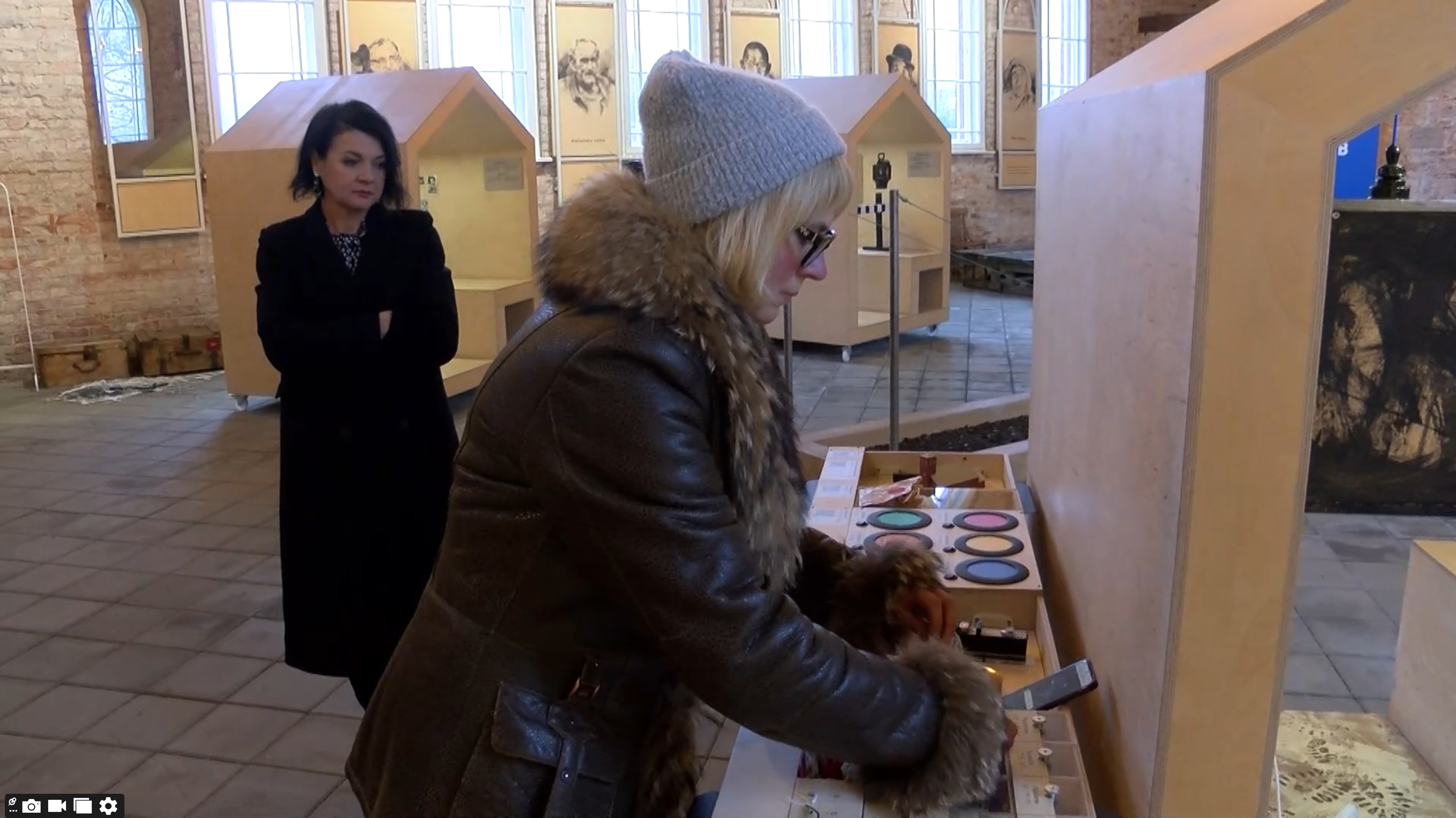 Sieviete cepurē un ziemas mētelī pēta taktilu ekspozīciju, kamēr fonā cita sieviete vēro notiekošo.