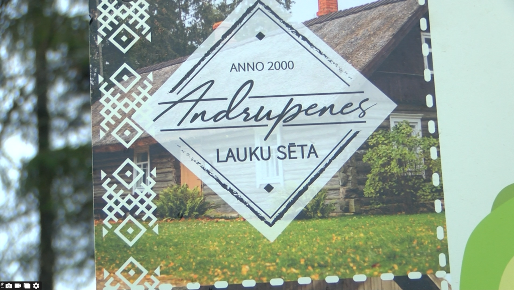 Tuvplāns informatīvai plāksnei ar uzrakstu "Andrupenes lauku sēta" un tradicionāliem latviešu rakstiem kreisajā malā.