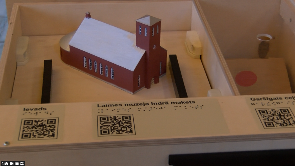 Sarkanbalts baznīcas ēkas makets koka kastē ar aprakstu Braila rakstā un QR kodiem priekšplānā.