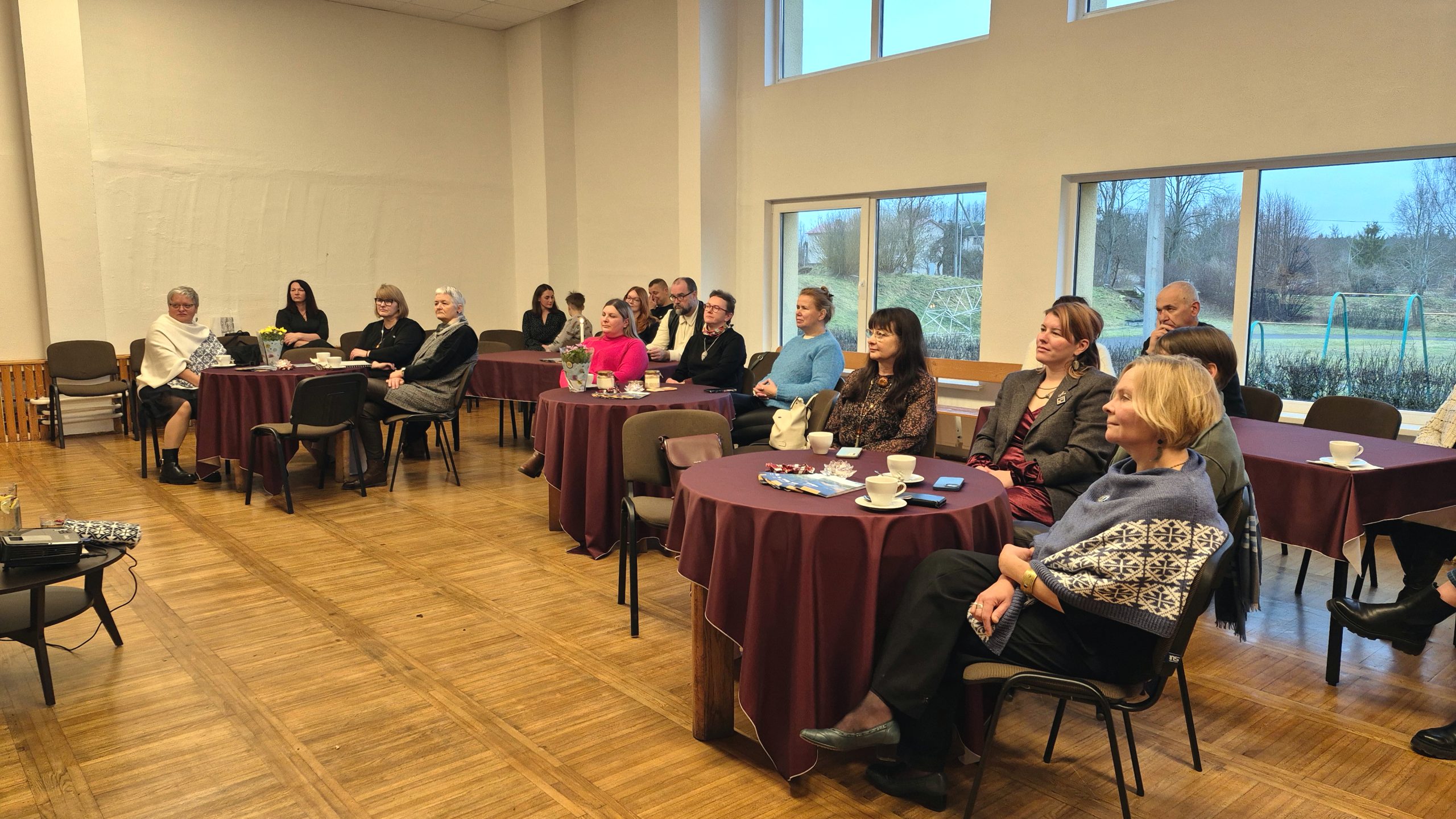 NVO pārstāvji un dalībnieki sēž pie maziem, apaļiem galdiem ar vīna sarkanām drānām kultūras projektu izvērtēšanas pasākumā Subatē.