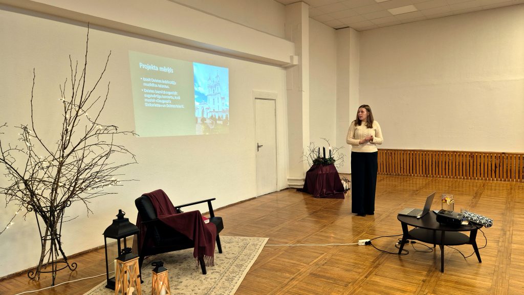 Sieviete stāv zālē pie sienas, uz kuras tiek projicēts slaids ar projekta mērķiem un Dvietes baznīcas attēlu.