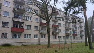 Daudzstāvu daudzdzīvokļu nams no baltiem ķieģeļiem ar lodžijām un blakus esošiem augstiem kokiem.