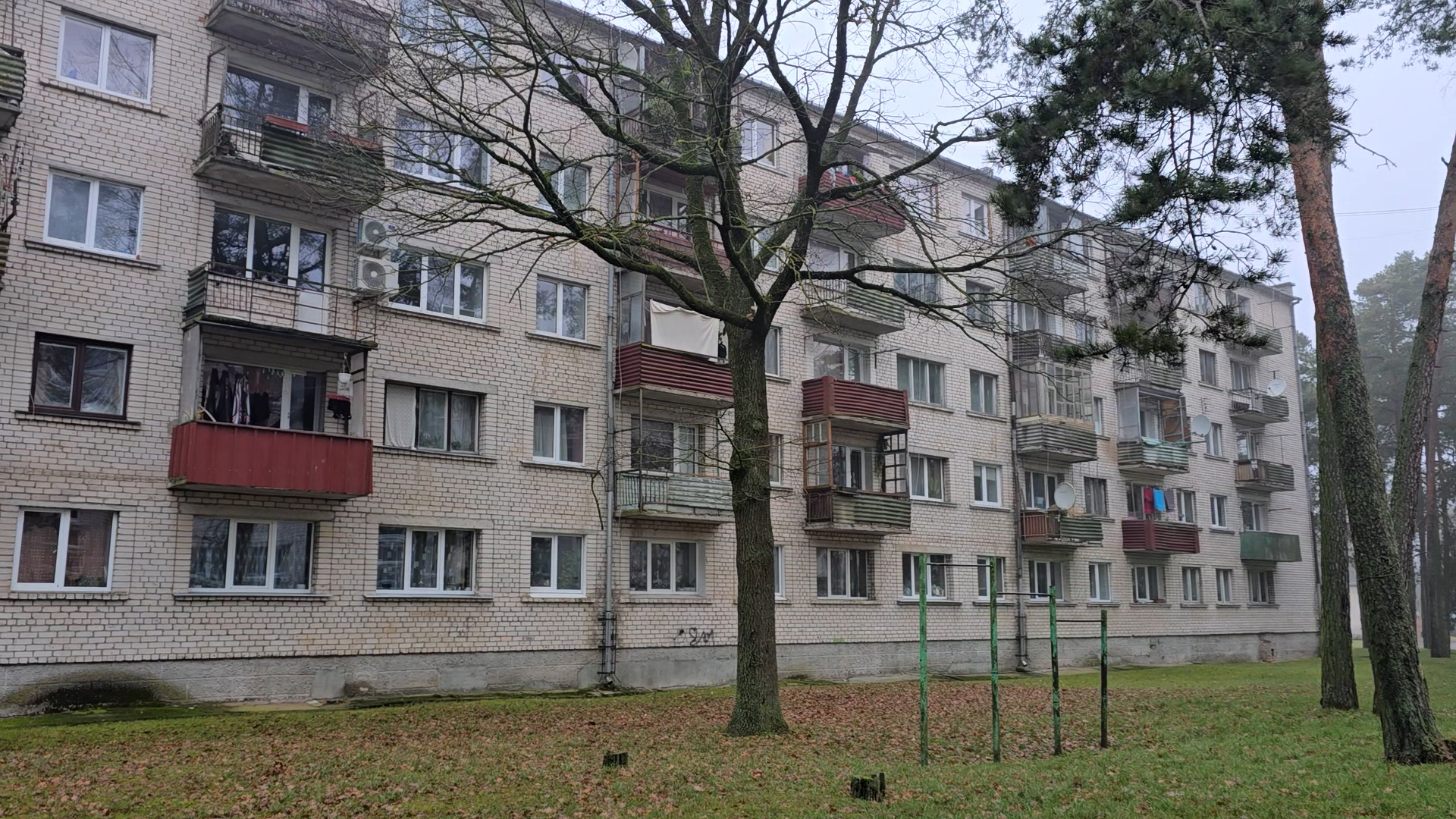 Daudzstāvu daudzdzīvokļu nams no baltiem ķieģeļiem ar lodžijām un blakus esošiem augstiem kokiem.