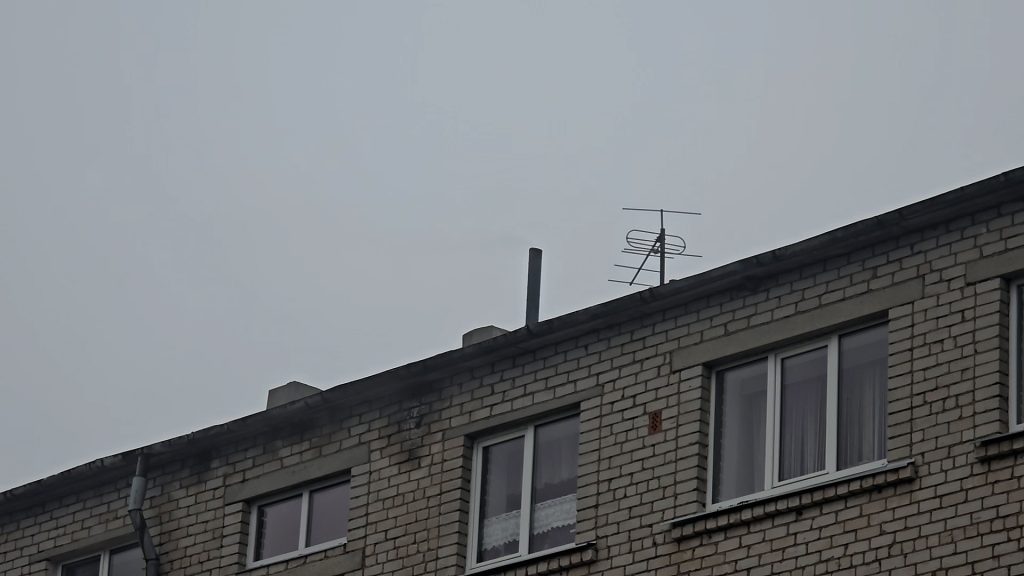 Tuvplāns uz daudzdzīvokļu nama jumta malu ar vienu nelielu melnu dūmeņu un antenu.