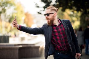 Jauns vīrietis ar modernu bārdu, saulesbrillēm un sarkanmelnu rūtainu kreklu, ārā turot telefonu un veidojot selfiju, izbāž mēli.