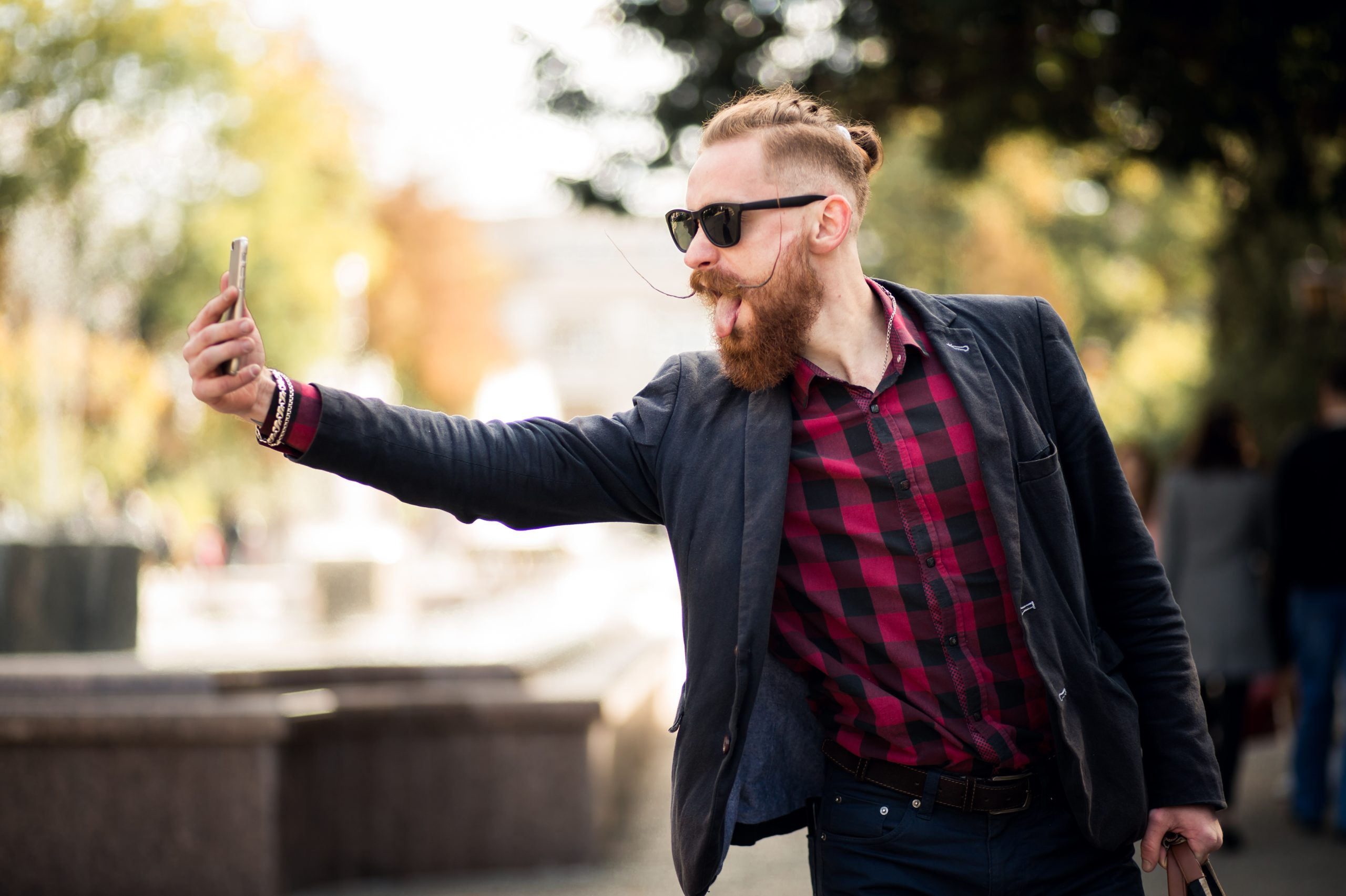 Jauns vīrietis ar modernu bārdu, saulesbrillēm un sarkanmelnu rūtainu kreklu, ārā turot telefonu un veidojot selfiju, izbāž mēli.