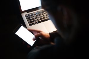 Persona tumsā lieto viedtālruni pie klēpjdatora.
