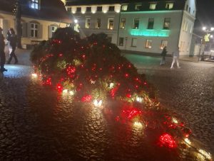 Vētrā apgāzta svētku egle Ventspils Rātslaukumā ar izgaismotām dekorācijām uz bruģa