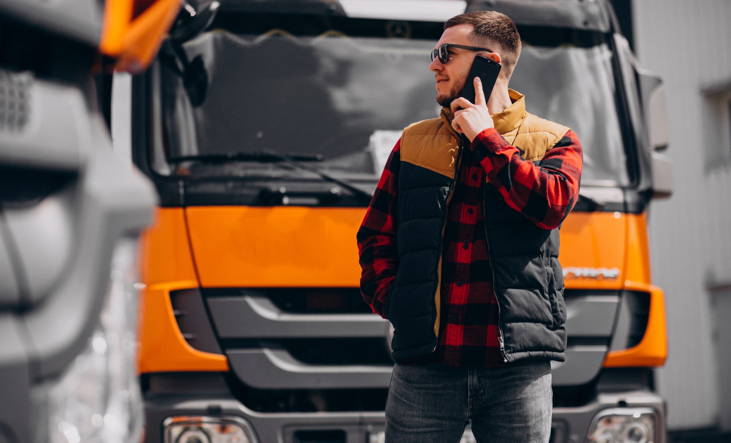 Kravas automašīnas vadītājs ar saulesbrillēm un džemperi stāv pie oranžas kravas automašīnas un runā pa mobilo telefonu.