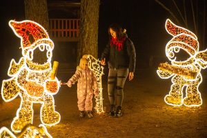Sieviete un bērns Tērvetes dabas parkā pie izgaismotām rūķu figūrām tumšā diennakts laikā.