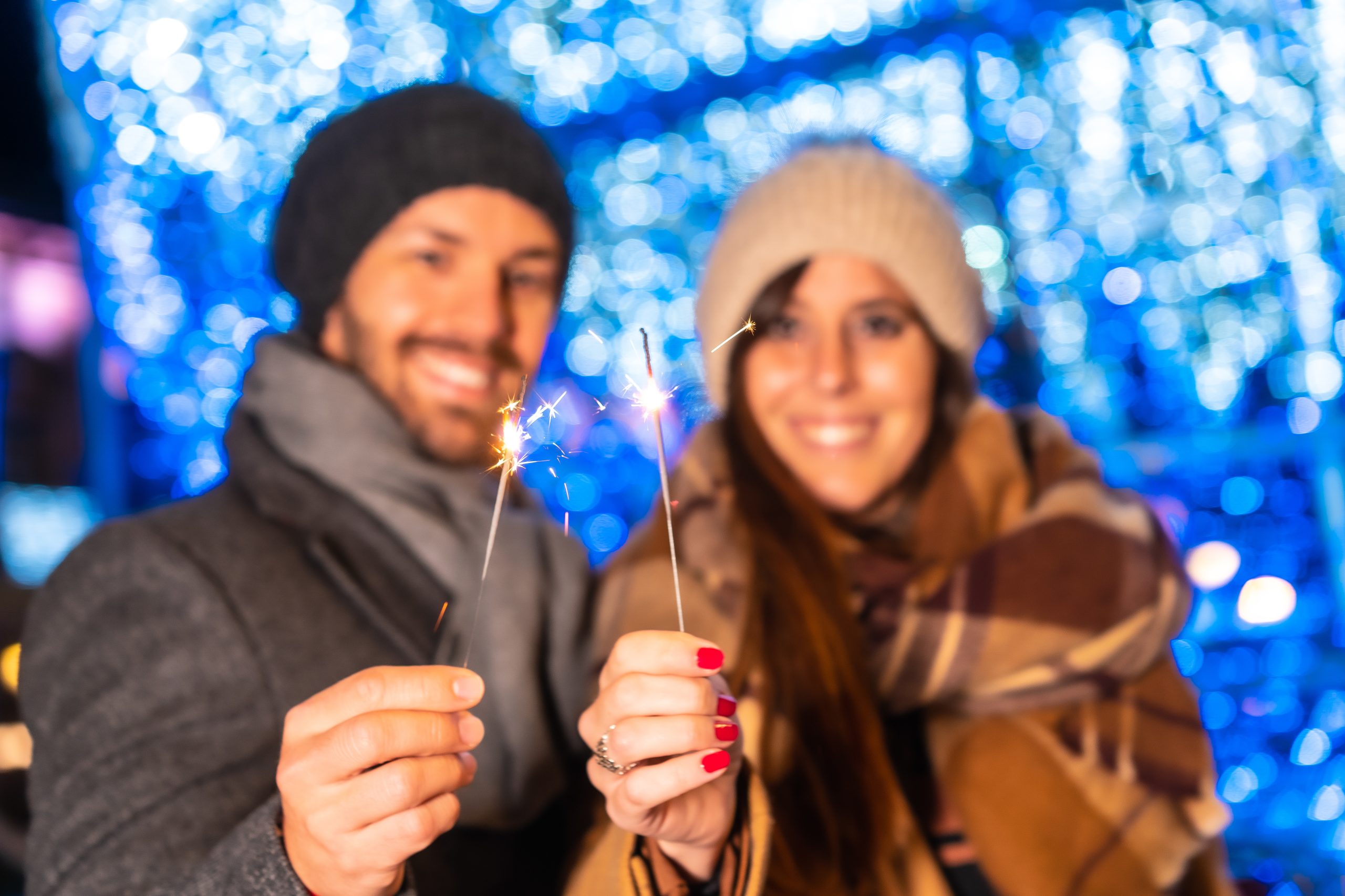 Smaidīgs pāris ziemas apģērbā tur rokās degošas brīnumsvecītes uz zila svētku apgaismojuma fona.