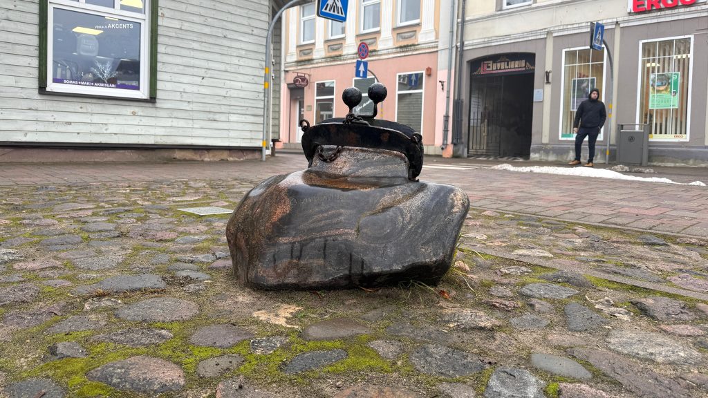 Skulptūra, kas atgādina tumšu, plakanu tēlu ar melnu maisiņu vai galvassegas virsmu ar diviem melniem apļiem, kas atgādina acis, uz veca bruģa.