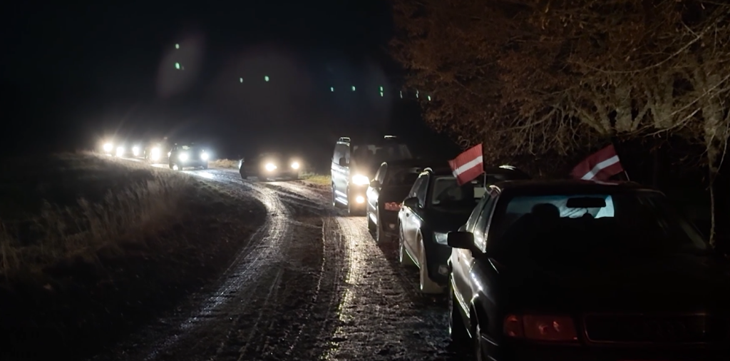 Automašīnu kolonna ar Latvijas karodziņiem brauc pa tumšu, dubļainu lauku ceļu.
