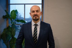 Vīrietis uzvalkā iekštelpās pie loga, ilustratīvs attēls rakstam par dezinformācijas un mākslīgā intelekta ietekmi