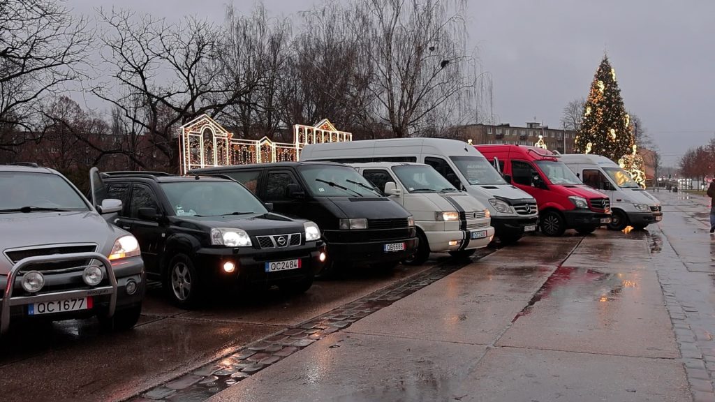 Rindā novietotas dažādas automašīnas, ieskaitot apvidus auto, mikroautobusus un mazus furgonus, lietainā dienā pilsētas laukumā pie izgaismotas egles.