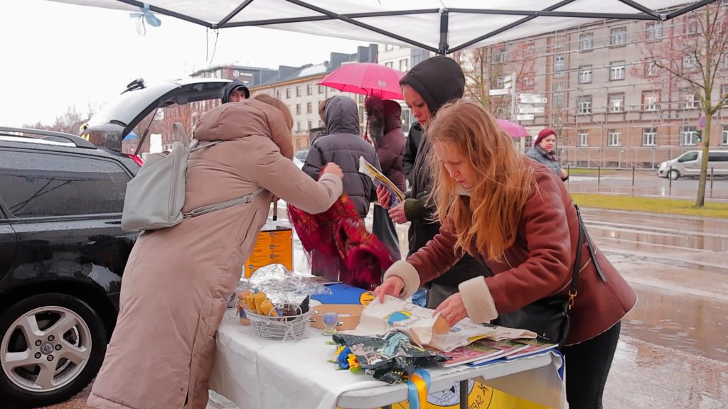 Cilvēki, vācot ziedojumus, strādā pie galda lietainā laikā zem nojumes, kraujot pārtiku un apģērbu automašīnā, kas tiks sūtīts uz Ukrainu.