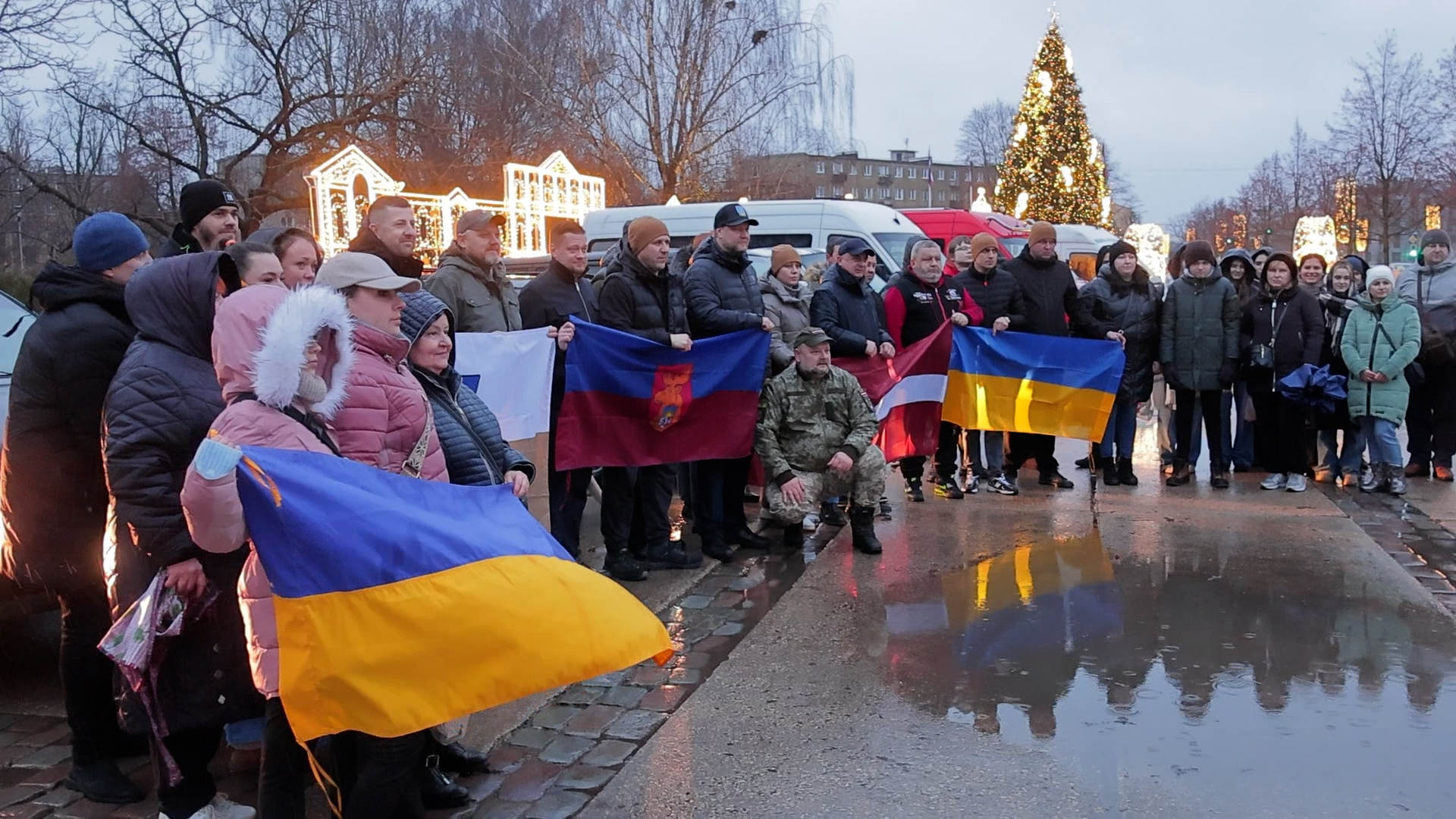 Cilvēku grupa stāv lietū pilsētas laukumā ar Latvijas un Ukrainas karogiem rokās, fonā izgaismota Ziemassvētku egle un dekorācijas.